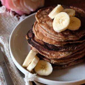 Stack of whole wheat banana pancakes topped with sliced bananas, maple syrup, and fresh berries, served with orange juice and soft pink flowers in the background.