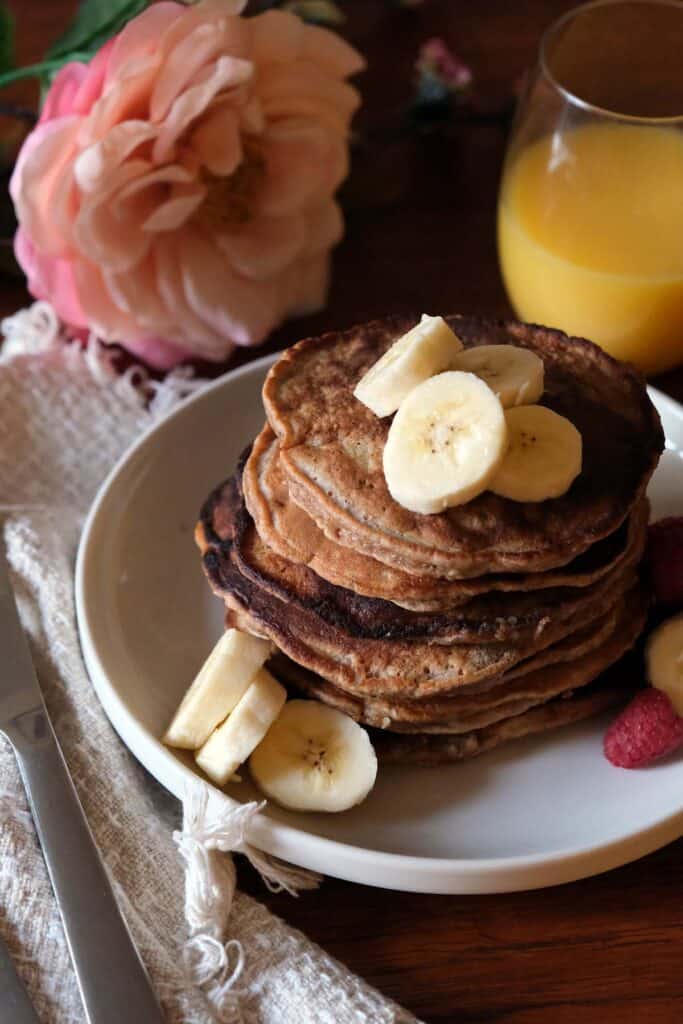 Stack of whole wheat banana pancakes topped with sliced bananas, maple syrup, and fresh berries, served with orange juice and soft pink flowers in the background.