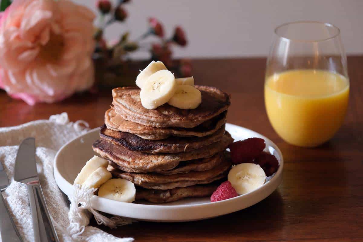 Healthy whole wheat banana pancakes stacked high with banana slices, berries, and maple syrup on a brunch table with orange juice and pink floral accents.