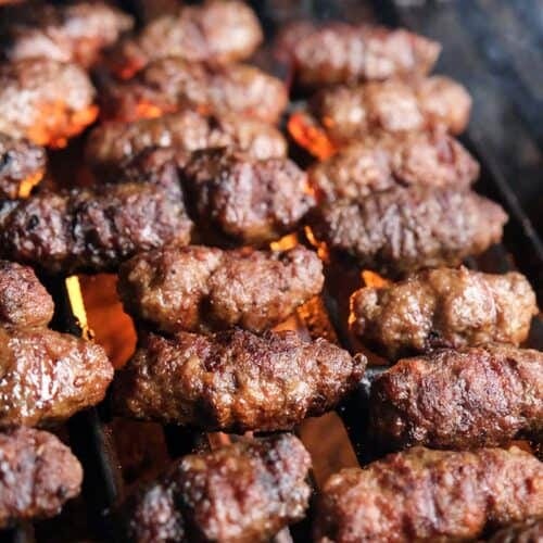 Ćevapčići grilling over open flame showing charred Balkan sausages cooking on barbecue