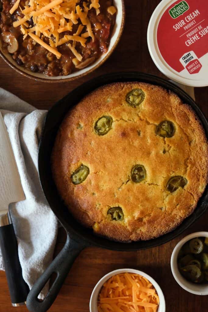 Top down view of skillet jalapeño cornbread in cast iron pan served with bowls of chili and toppings