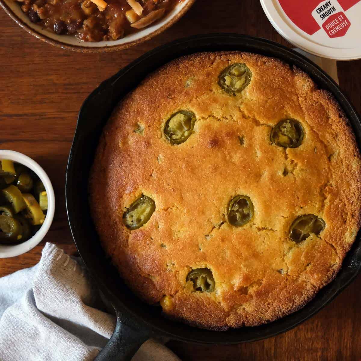Cast iron skillet jalapeño cornbread surrounded by chili bowls, sour cream, and toppings