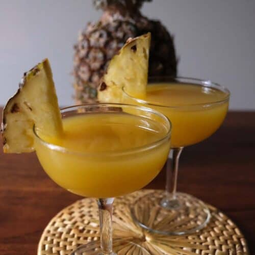 Pineapple Coconut Rum Cocktail in coupe glasses garnished with fresh pineapple slices, with a whole pineapple in the background